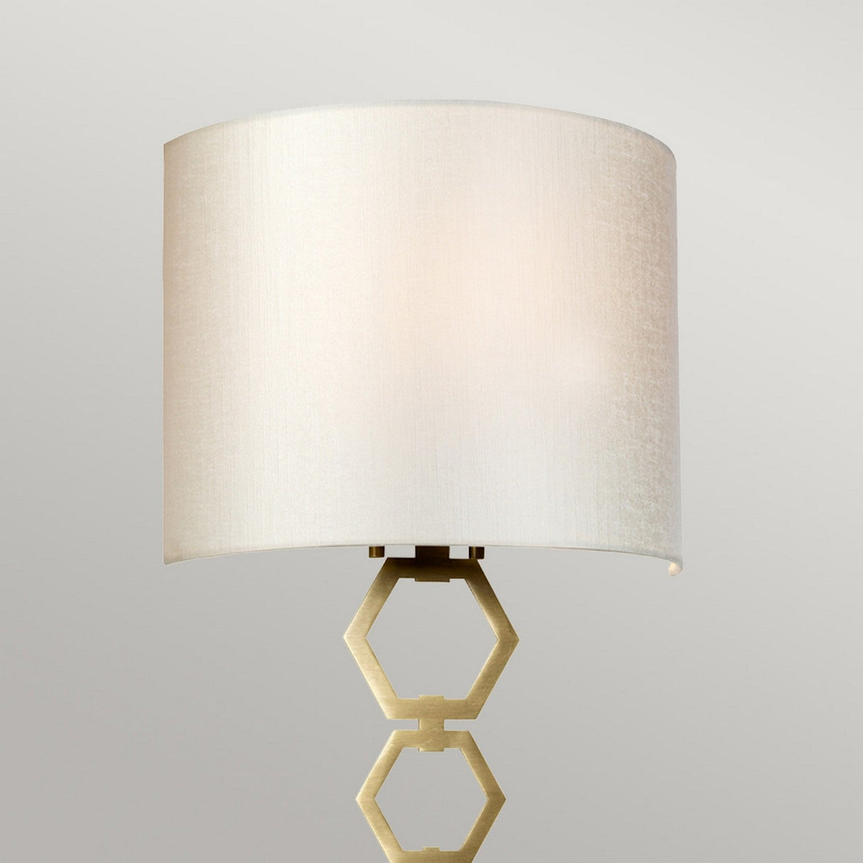 The Clark Medium Wall Light in aged brass is a contemporary lighting fixture showcasing a faux silk shade with an off-white cylindrical design, elegantly paired with a geometric hexagonal metal stand, creating an ideal atmosphere against a neutral background.