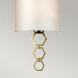 A close-up of the Clark Medium Wall Light - Aged Brass reveals three interlocking hexagonal rings elegantly suspended below a faux silk shade. The sleek and minimalist design of this ambient lighting piece stands out against a plain background.