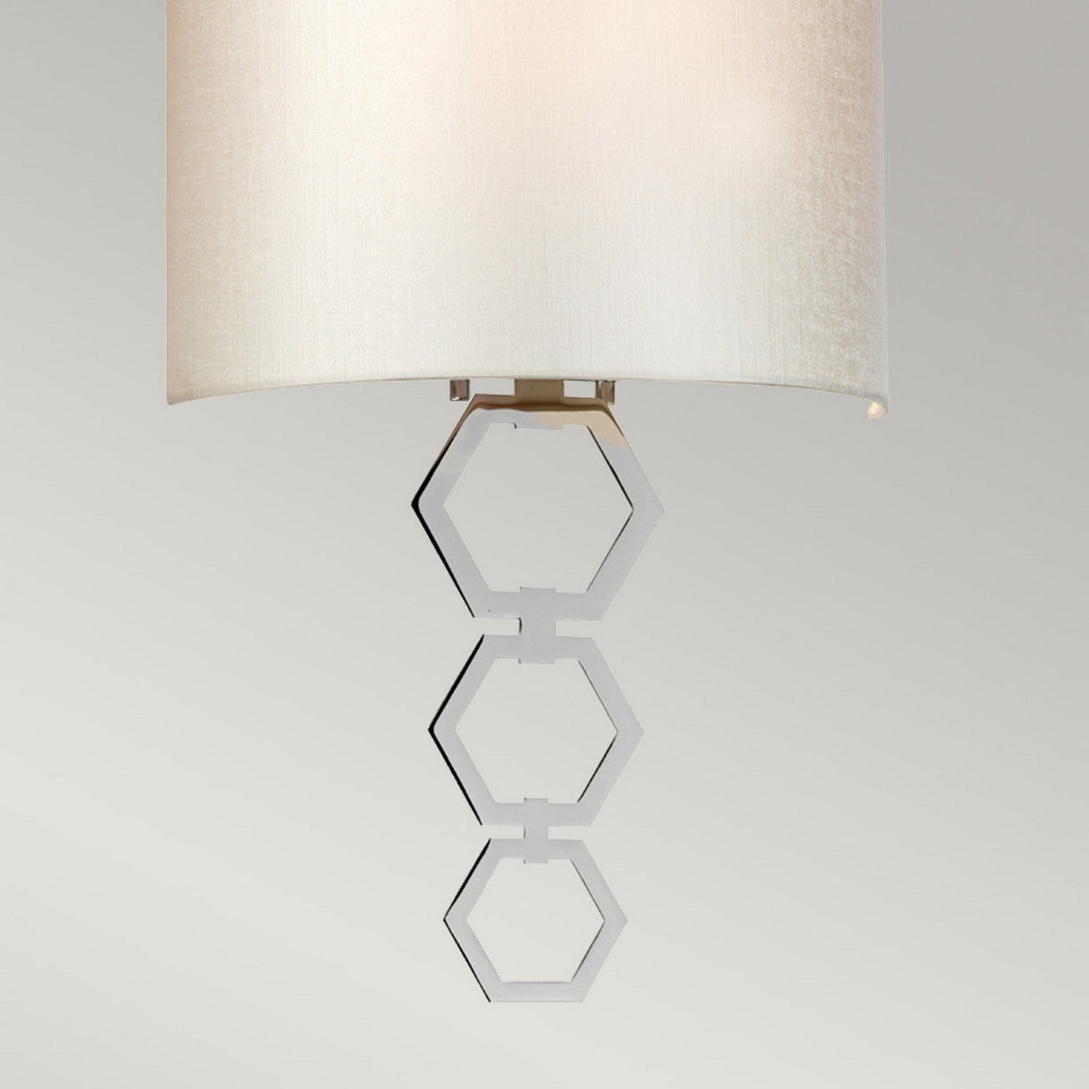 The Clark Medium Wall Light in polished chrome showcases a contemporary design with a faux silk beige shade and an elegant decorative pull chain featuring three interconnected hexagonal rings in a metallic finish, ideal for modern interiors. The background is a subtle, soft gray.