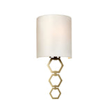 The Clark Small Wall Light - Aged Brass showcases a contemporary design with a white faux silk shade, enhanced by a geometric hexagonal pattern base in aged brass, stylishly installed on a white wall.