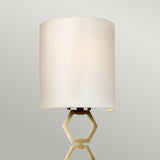 A contemporary wall light featuring a beige fabric shade and a geometric, aged brass base set against a light gray backdrop.