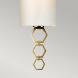 The Clark Small Wall Light - Aged Brass boasts a modern beige sconce with a geometric hexagonal design, complemented by an aged brass fixture. Its cylindrical faux silk shade elegantly contrasts with the neutral background.