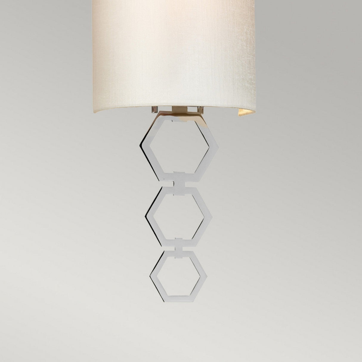 The Clark Small Wall Light - Polished Chrome features a contemporary light fixture with a faux silk cylindrical white shade, embellished with three polished chrome hexagon rings hanging from its base, all set against a plain gray background.