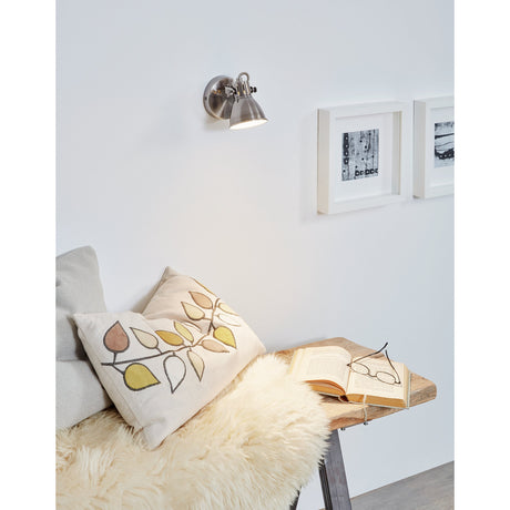 Clasanne wall spotlight in antique nickel steel with cream-lined bell shade, mounted above a reading nook with cushions and framed artwork.