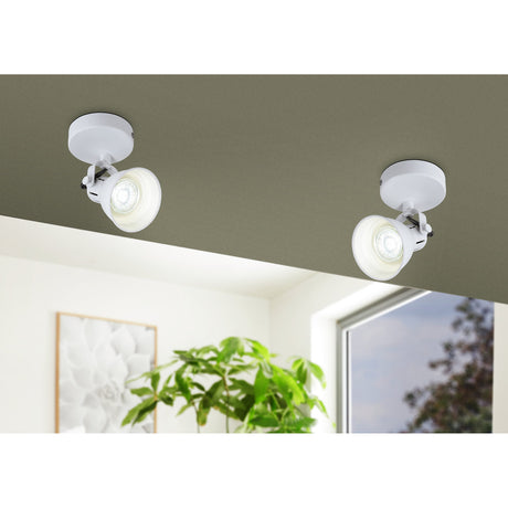 Two Clasanne single-head wall/ceiling spotlights in white steel with round mounting plates, mounted on a grey ceiling edge above a bright room with a plant and framed art.