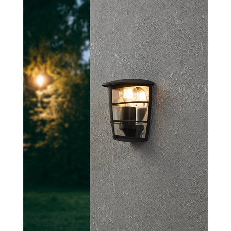 Clasvarne black outdoor wall light with half-lantern design and clear panels, mounted on a textured grey exterior wall, lit E27 bulb visible inside.