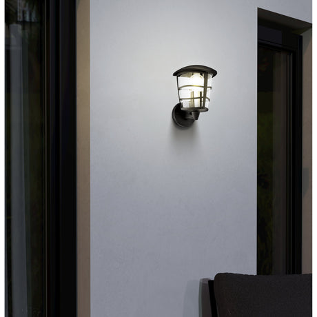 Clasvarne upward-facing outdoor wall light in black cast aluminium with a clear lantern-style shade, illuminated on an exterior wall beside a doorway.