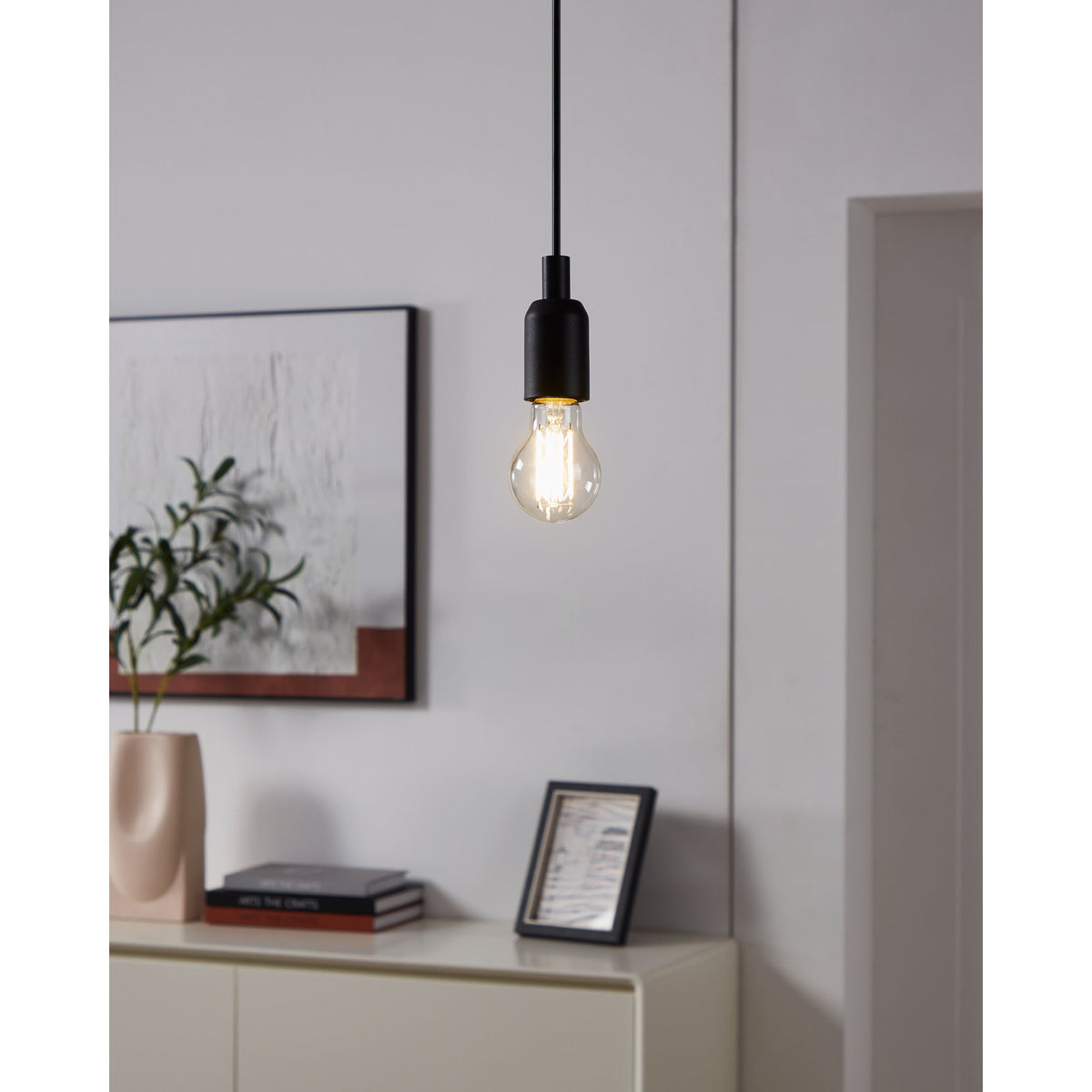 Clear GLS LED filament bulb with E27 cap glowing warm white on a black pendant cord above a sideboard in a living room setting.