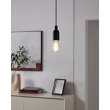 Clear GLS LED filament bulb with E27 cap glowing warm white on a black pendant cord above a sideboard in a living room setting.