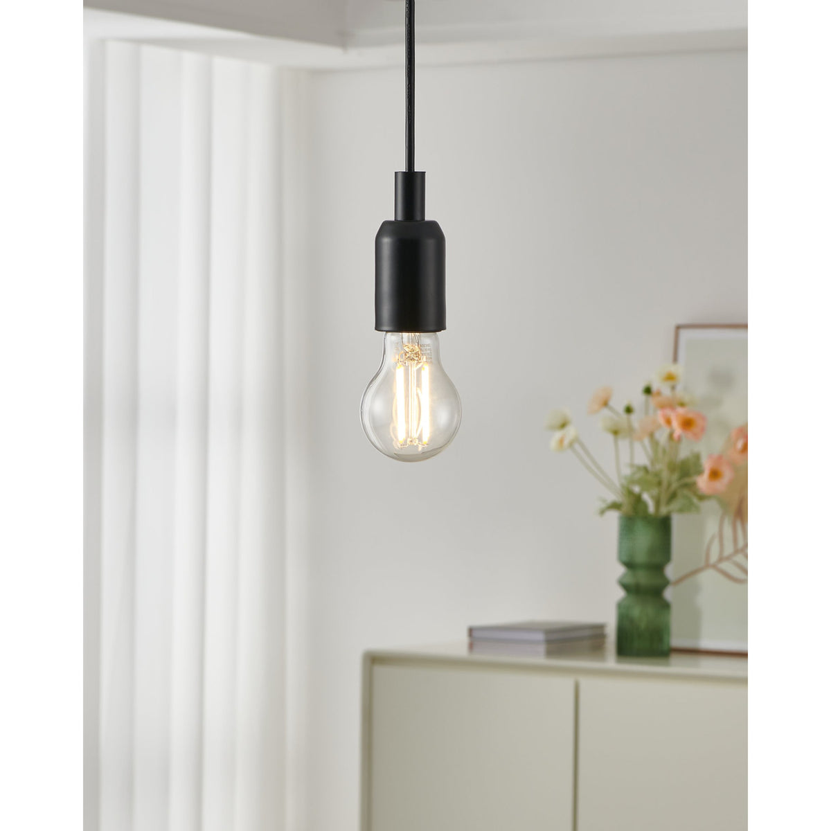 Clear LED Bulb E27 7W 806lm CCT with visible upright filaments, shown glowing warmly in a matte black pendant fitting above a light-filled room with flowers and a sideboard.