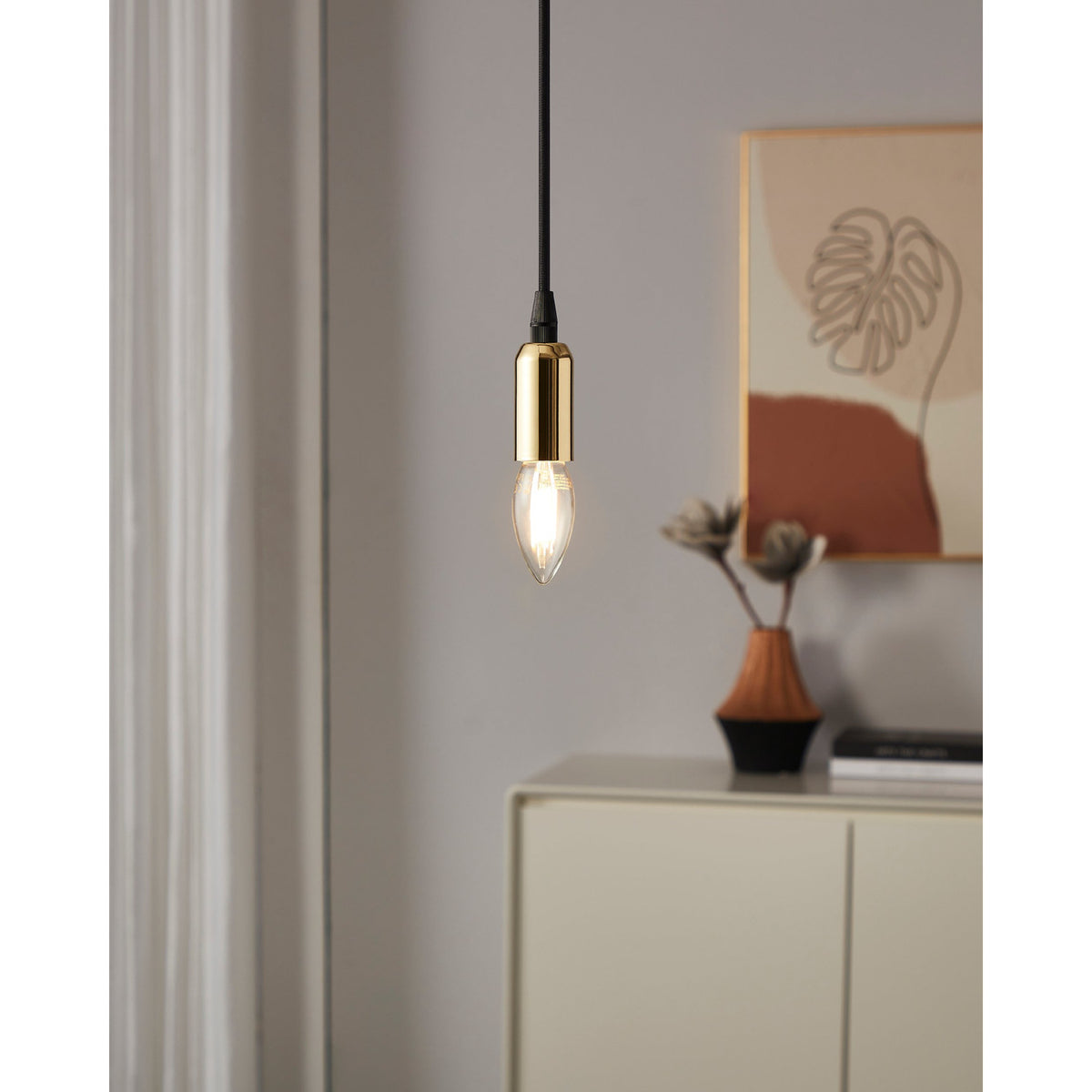 Clear LED candle bulb E14 with visible filament strips, shown glowing warm white in a brass pendant light fitting above a cream sideboard.