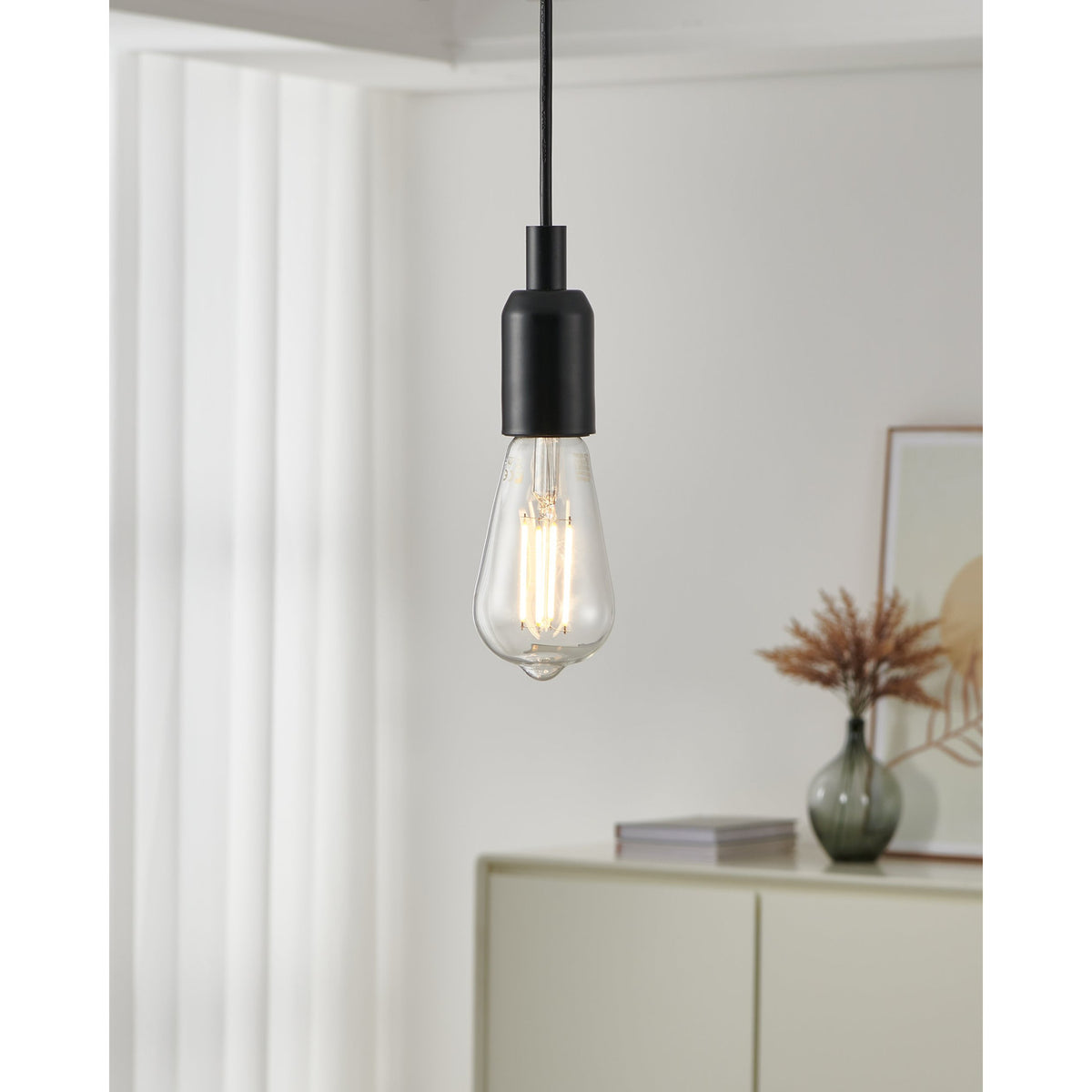 Clear LED Vintage Bulb E27 ST64 teardrop shape with visible filaments, glowing warm white in a matte black pendant fitting above a light-coloured sideboard.