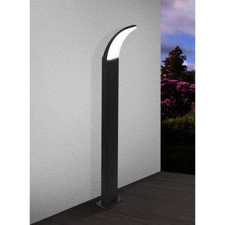 Clesmane LED Bollard Light 90cm in black, a slim square aluminium post with curved head and white diffuser, shown illuminated against an outdoor wall with decking and garden planting.