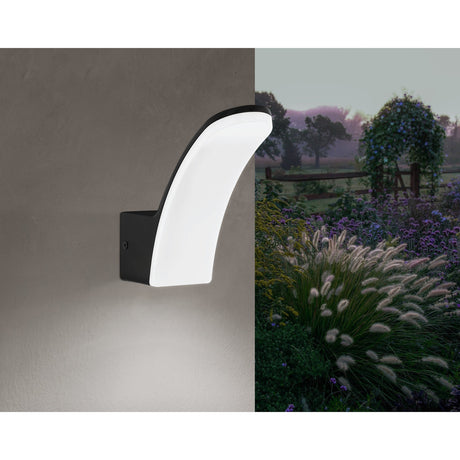 Clesmane LED Outdoor Wall Light in black, with a curved aluminium frame and white plastic diffuser, shown illuminating a wall beside a garden setting at dusk.