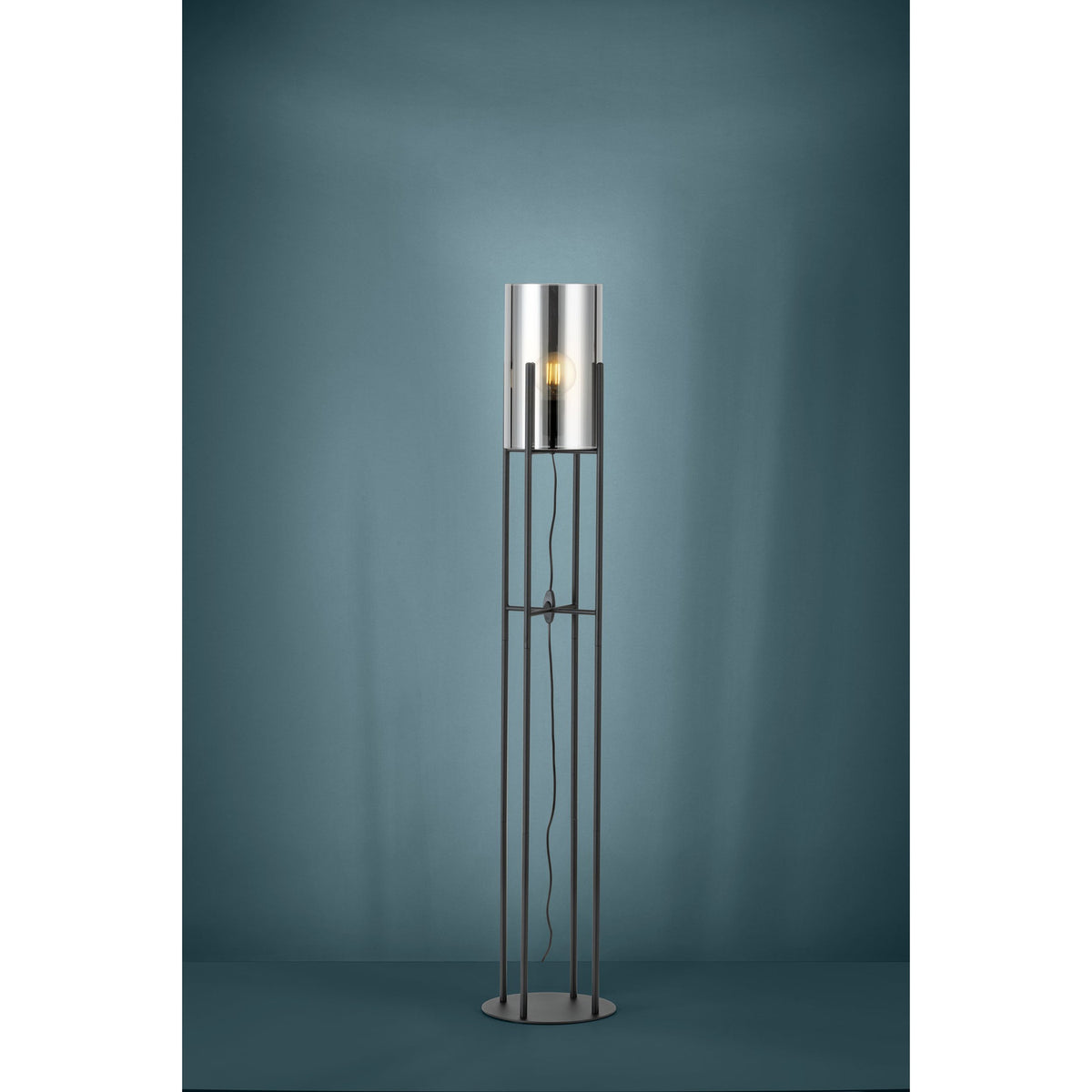 Clisvelle Floor Lamp in black steel, featuring four slim upright rods on a round base supporting a cylindrical vaporised glass shade with a visible E27 bulb, shown against a teal studio background.