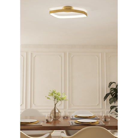 Clomelle LED flush ceiling light in brushed brass, a rounded square ring with white diffuser, shown illuminated above a panelled dining room with wood table and gold-accented plates.
