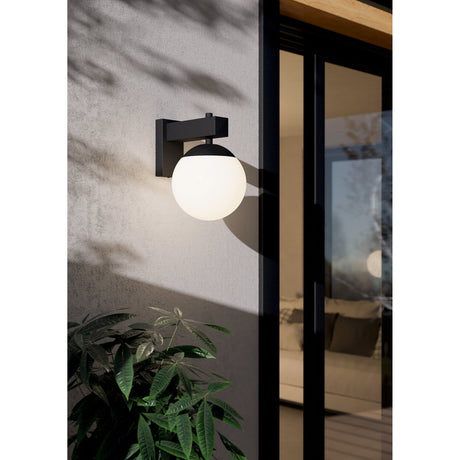 Cloressa outdoor wall light in black, with a square steel bracket holding a white globe shade, illuminated beside a glass door on a textured grey exterior wall.