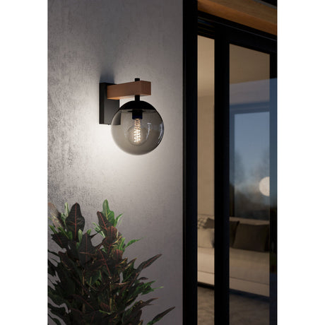 Cloressa outdoor wall light with black steel backplate, wood-grain brown arm and smoked globe shade, illuminated beside a glass door at night.
