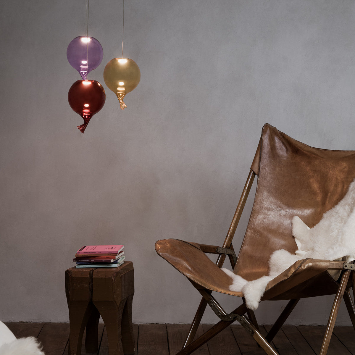 Clown 15cm Glass Light Shade in red, a balloon-shaped pendant with twisted glass tail, shown alongside purple and amber versions above a leather chair and wooden stool.