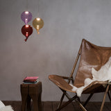 Clown 15cm Glass Light Shade in red, a balloon-shaped pendant with twisted glass tail, shown alongside purple and amber versions above a leather chair and wooden stool.
