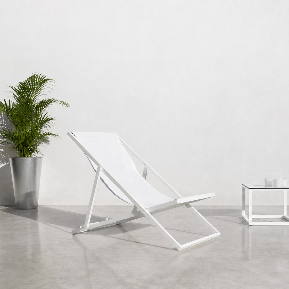 Club Aluminium Folding Deck Chair in white, reclined sling seat on an aluminium frame, shown beside a potted palm and glass side table on a light concrete floor.