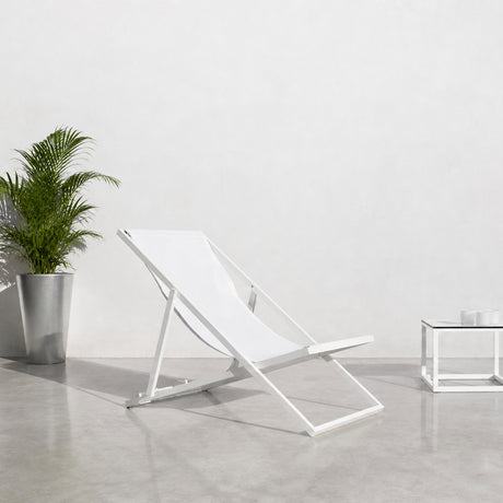 Club Aluminium Folding Deck Chair in white, reclined sling seat on an aluminium frame, shown beside a potted palm and glass side table on a light concrete floor.