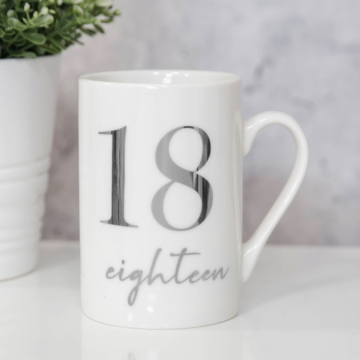 White ceramic 11oz mug with a large silver foil "18" and "eighteen" in script below, shown with a plant against a neutral background.