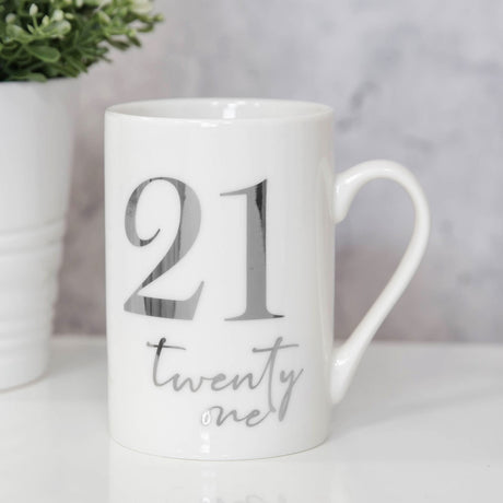 White ceramic mug with "21 twenty one" in silver foil lettering, tall straight shape with curved handle, shown on a table with a plant in the background.