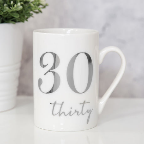 White ceramic 11oz mug with silver foil "30" and "thirty" script, curved handle, shown beside a potted plant on a light surface.