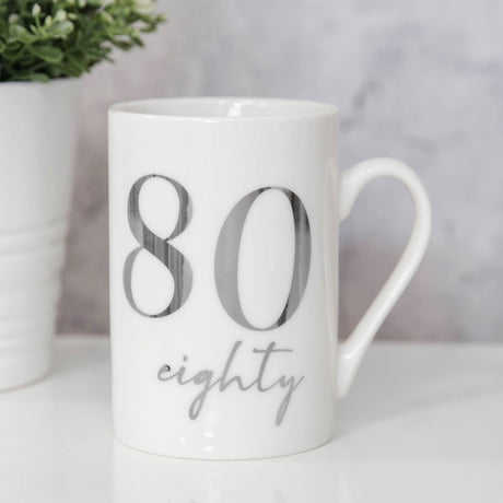 White ceramic birthday mug with "80" in bold silver foil and "eighty" in silver script beneath, glossy finish, shown on a table with a small plant in the background.