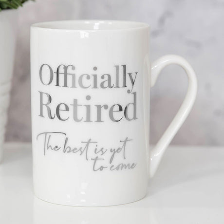 White ceramic mug with silver foiled text "Officially Retired, The best is yet to come," straight sides, open handle, shown on a light table setting.