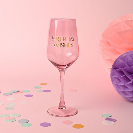 Pink stem wine glass with "BIRTHDAY WISHES" in gold lettering on the bowl, slender stem and round foot, set against a pink background with confetti and tissue paper pom-poms.