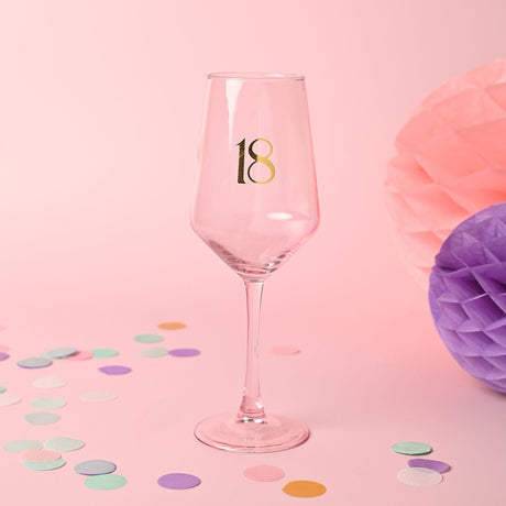 Pink glass wine glass with a gold "18" printed on the bowl, tall slim stem and round foot, set against a pink background with confetti and tissue paper pom-poms.