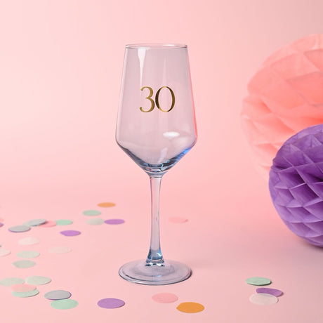 Light blue wine glass with gold "30" numeral on the bowl, slim stem and round foot, set against a pink background with confetti and tissue paper pom-poms.