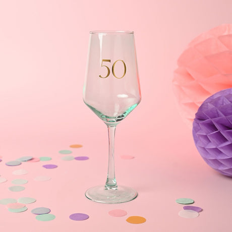 Stemmed wine glass with pale green tint and gold "50" on the bowl, slim stem and round foot, set against a pink background with confetti and pom-poms.