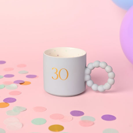 Blue porcelain mug with gold "30" numeral, matt finish, bobble ring handle, and star-patterned cream interior, shown on a pink background with confetti.