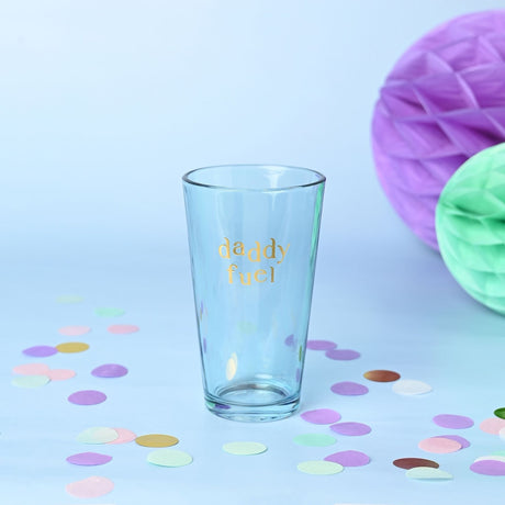Clear glass beer tumbler with "daddy fuel" in gold foiled lettering, tapered pub-glass shape, set against a light blue background with pastel confetti and honeycomb decorations.