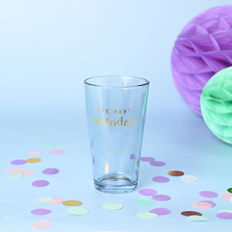 Clear tapered beer glass with "THE BEST Grandad" in gold foil lettering, set on a light blue background with pastel confetti and honeycomb decorations.