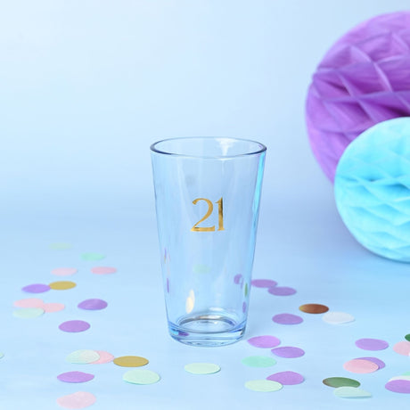 Clear tapered beer glass, 460ml, with gold foiled number 21 on the front, set against a light blue background with pastel confetti and paper decorations.