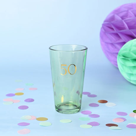 Green-tinted tapered beer glass with gold foil "50" in serif font, 460ml capacity, shown on a light blue background with pastel confetti and honeycomb decorations.