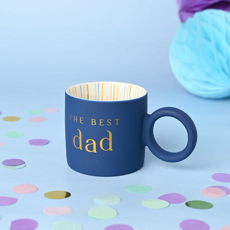 Navy porcelain mug with "THE BEST dad" in gold lettering, full round ring handle, and striped cream interior, set against a light blue background with confetti.