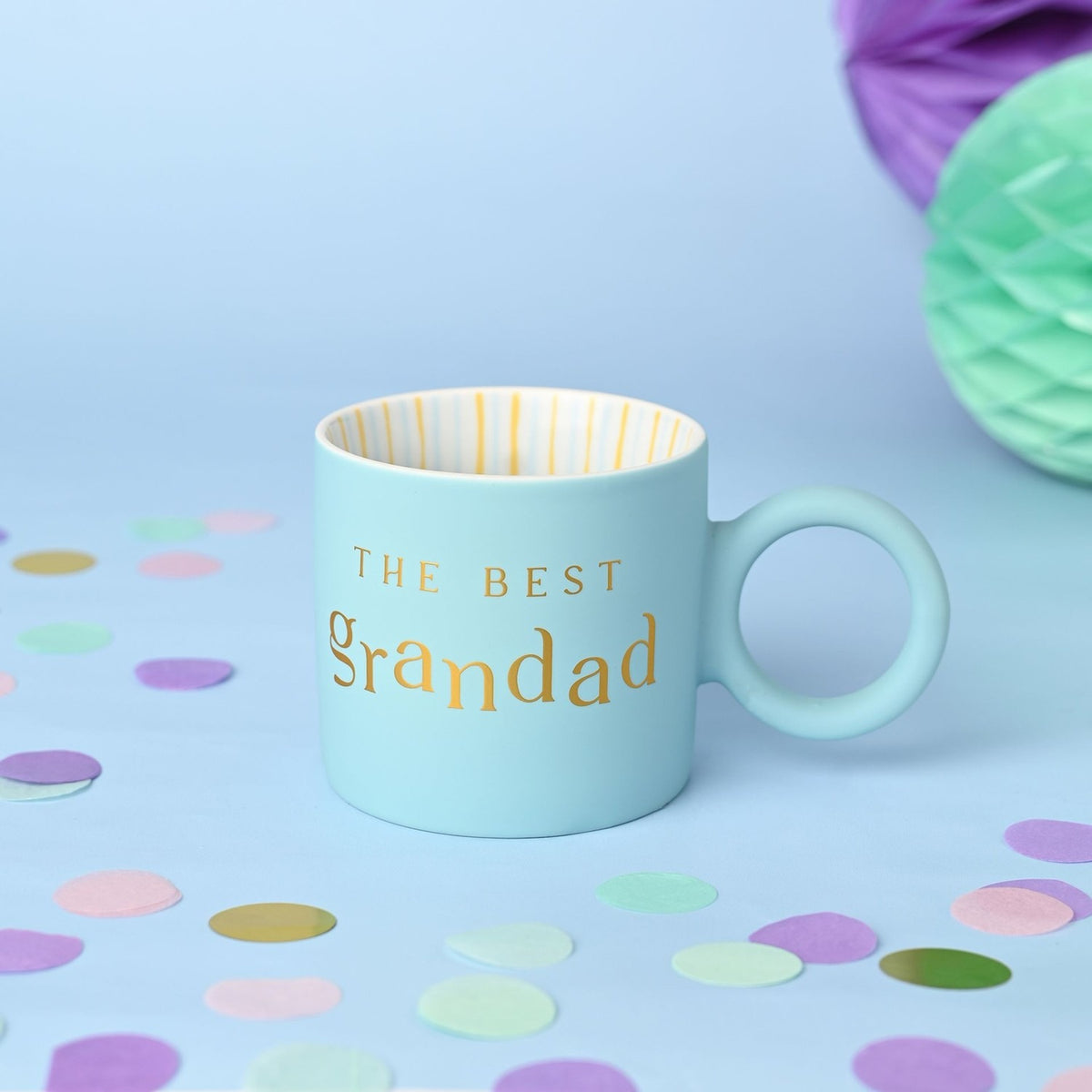 Light blue matt porcelain mug with "THE BEST grandad" in gold lettering, gold-striped white interior, shown with pastel confetti and paper honeycomb decorations.