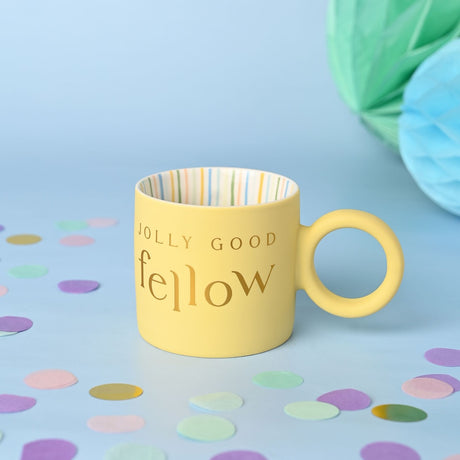 Yellow porcelain mug with round ring handle and gold "JOLLY GOOD fellow" text, striped interior, set on a light blue background with confetti.