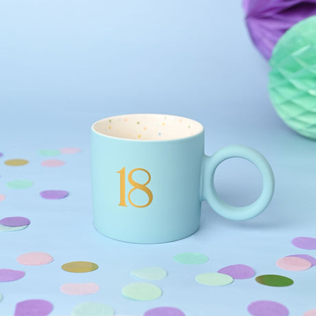 Light blue matt porcelain mug with gold "18" numeral and round ring handle, cream interior speckled with multicoloured dots, shown with pastel confetti and paper decorations.