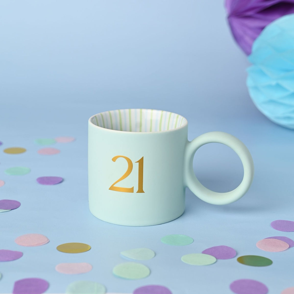 Light blue matt porcelain mug with a round ring handle, featuring a gold "21" on the front and striped green and pale blue interior, set against a light blue background with confetti.