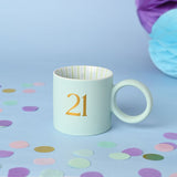Light blue matt porcelain mug with a round ring handle, featuring a gold "21" on the front and striped green and pale blue interior, set against a light blue background with confetti.