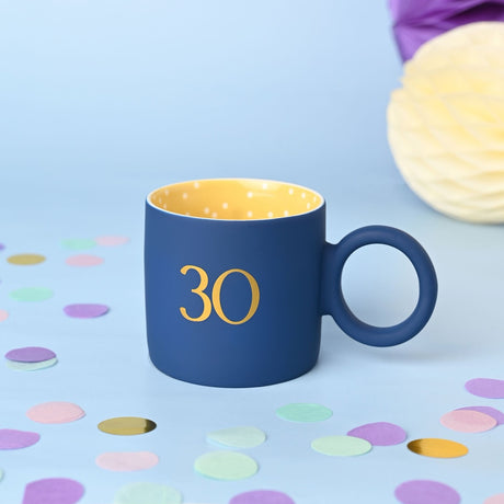 Navy matt porcelain mug with gold "30" numeral, yellow polka-dot interior and round ring handle, shown against a light blue background with confetti.
