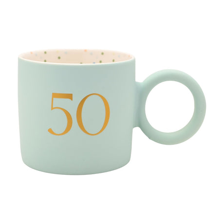 Light blue matt porcelain mug with gold "50" text and a large round ring handle, cream interior with multicoloured dots, shown on a plain white background.