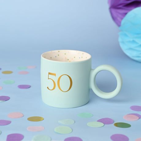 Light blue matt porcelain mug with a gold "50" and round ring handle, speckled cream interior, shown on a light blue surface with scattered confetti.