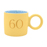 Matt porcelain mug in pastel yellow with a gold "60" print, blue polka-dot interior, and a chunky round ring handle, shown on a plain white background.